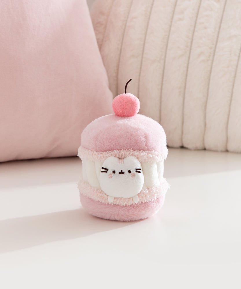 Cat plush toy resembling a macaron with a cherry on top, sitting on a light pink surface. A white Pusheen the Cat mini plush is able to removed from the macaron plush environment. 