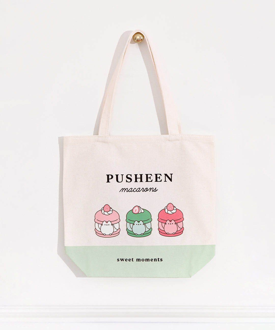 The white canvas bag with pink, green, white, grey, and brown printed graphic hangs on a gold knob in front of a white background. 
