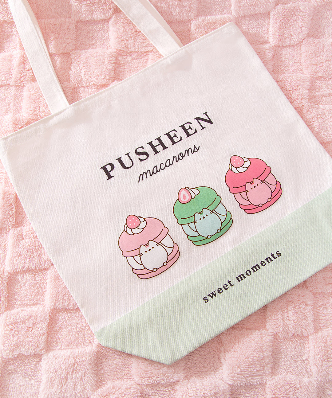 Close-up view of the printed graphic on the front of the Pusheen Sweets Tote Bag. The grey and brown tabby cat is inside three different macarons that are vanilla, matcha, and strawberry flavored.