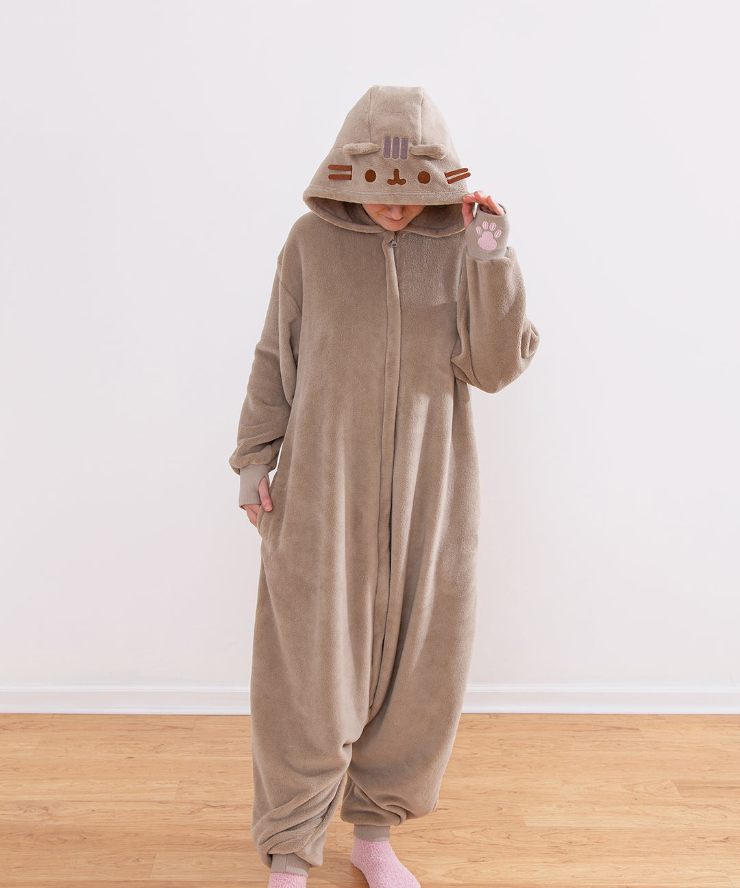 Model wearing a brown Pusheen Toe Beans Kigurumi onesie with a hood that has Pusheen the Cat's face embroidered on it and has 3D brown whiskers.