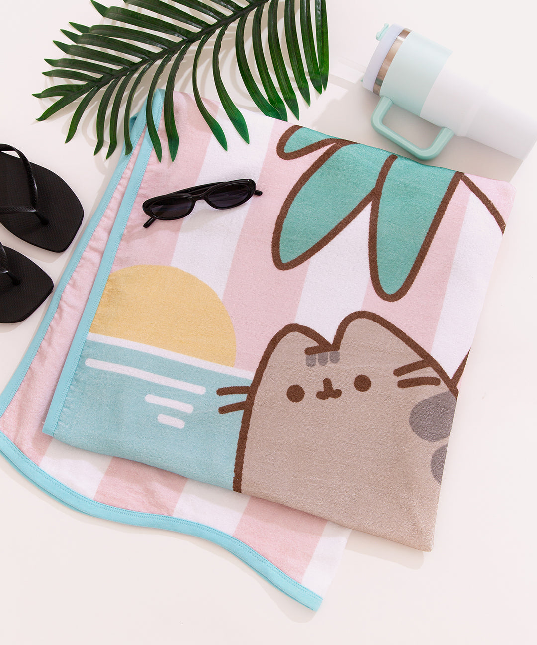 Beach towel with Pusheen graphic shown with pool day accessories including sandals, sun glasses, and a water bottle.