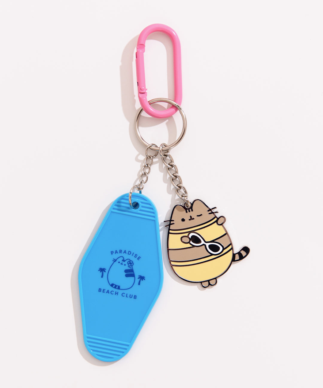 Close-up of the Pusheen Tropical Vacation Keychain that has a blue plastic keychain that says "paradise beach club" and a metal keychain of Pusheen winking while wearing a yellow bikini and holding sunglasses. The two keychains are attached to a silver split ring and pink carabiner.