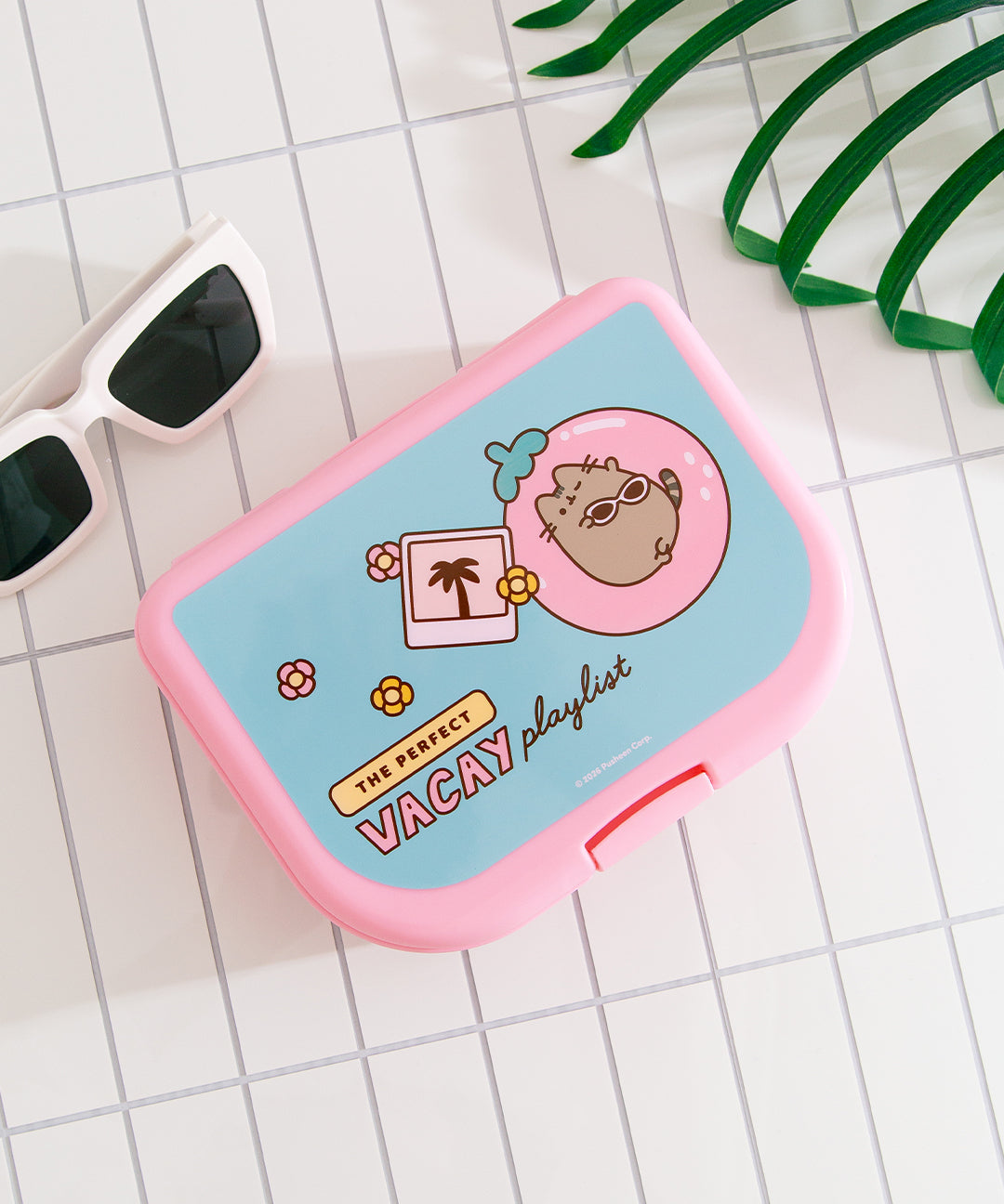 Tropical Vacation Pusheen Lunch Box lies on a table top. The pink box has a secure clasp on the side to keep the box together. The top of the box has a blue, pink, and yellow graphic of Pusheen.