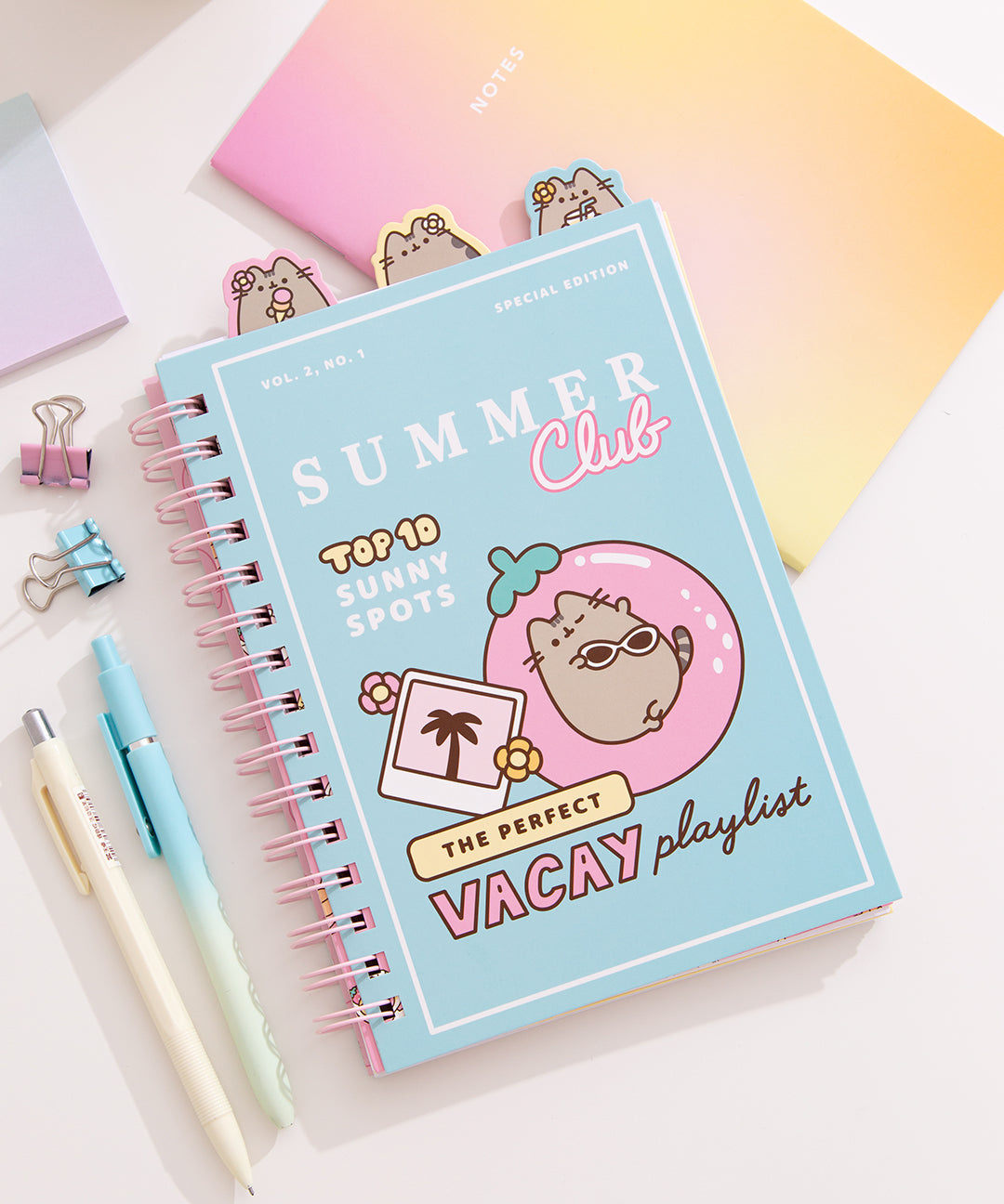 A light blue spiral bound notebook features graphcs of Pusheen in a strawberry pool float surrounded tropical vacation phrases including "summer club," "The perfect vacay playlist," and "top 10 sunny spots."