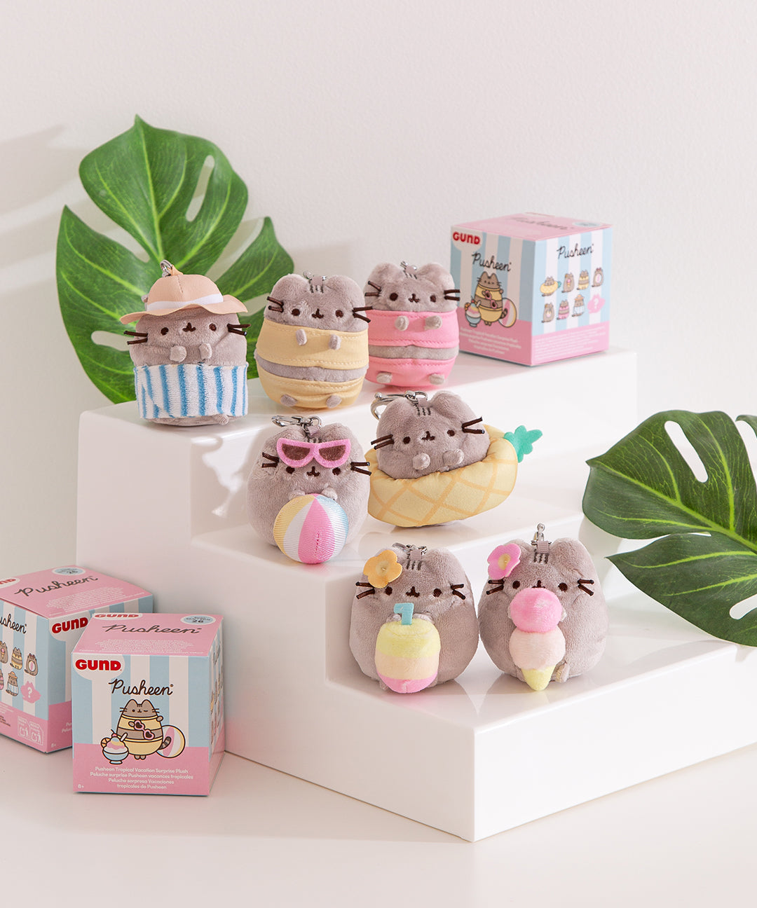 Front view of Pusheen Tropical Vacation Surprise Plush assortment. Plush keychains sit on multi-level white pedestal in front of a desktop background. Pusheen wears various poolside looks and holds summery snacks.