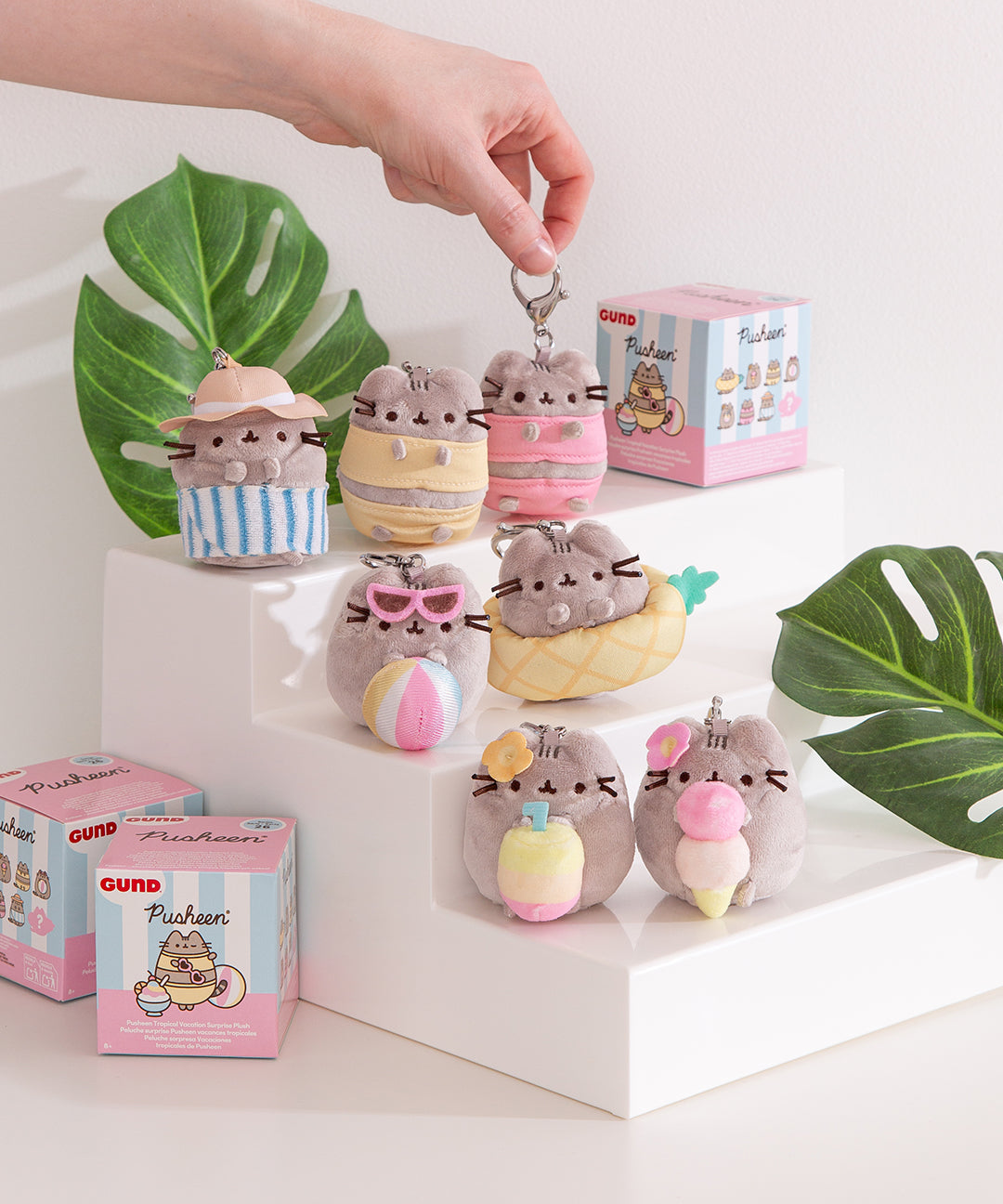 Pusheen Tropical Vacation Plush blind box selection sits on a white pedestal and does not show the hidden chase plush. A model holds the keychain attached to the top of the plush to show that each plush comes with a lobster clasp.