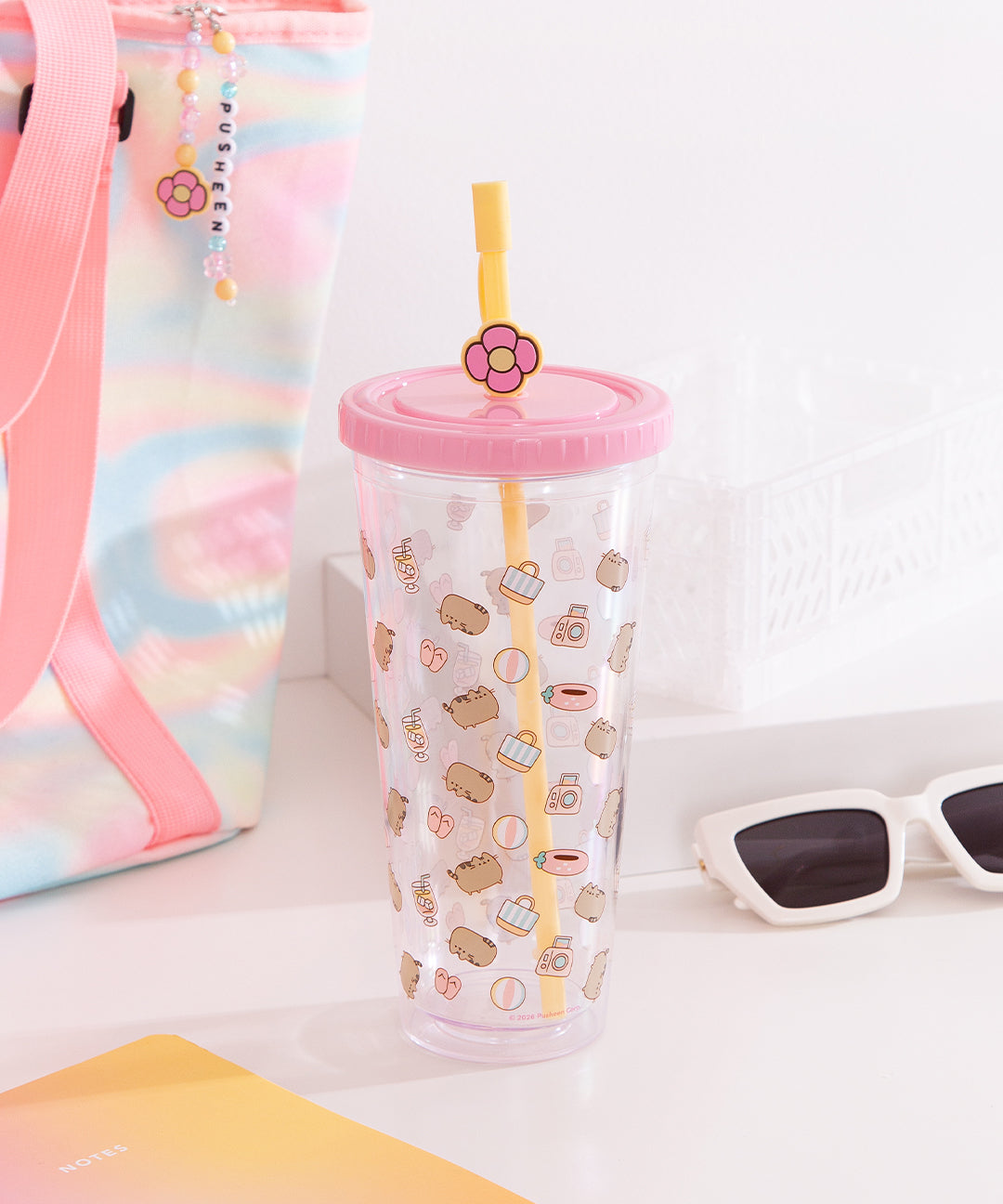 Front view of the Pusheen Tropical Vacation Tumbler. The clear beaker has a repeating pattern shows Pusheen surrounded by beach tote bags, beach balls, strawberry floaties, sandals, tropical drinks, and cameras.