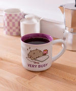 Mug with printed Pusheen graphic on each side. The white mug is placed in front of a kettle and spoon.