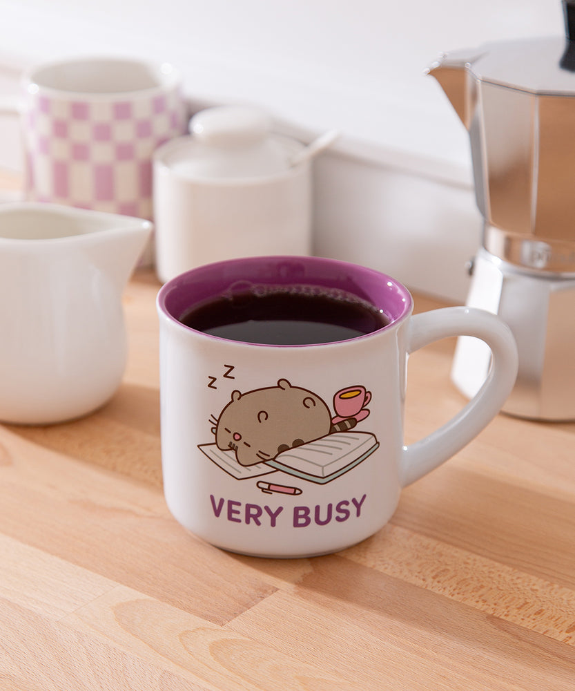 Pusheen Very Busy Mug is white with a purple interior and features Pusheen the Cat sleeping on papers.