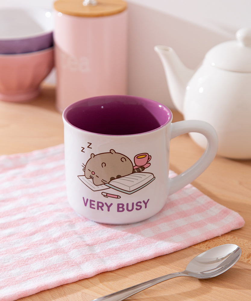 Mug with printed Pusheen graphic on each side. The white mug is placed in front of a kettle and spoon.