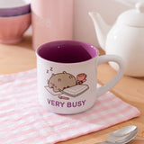 Mug with printed Pusheen graphic on each side. The white mug is placed in front of a kettle and spoon.