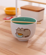 Pusheen Yum Ramen Bowl is a ceramic bowl with a white exterior and teal interior. One side of the bowl features a Pusheen the Cat graphic. The included chopsticks are shown lying in the notch at the top of the bowl. 