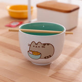 Pusheen Yum Ramen Bowl is a ceramic bowl with a white exterior and teal interior. One side of the bowl features a Pusheen the Cat graphic. The included chopsticks are shown lying in the notch at the top of the bowl. 