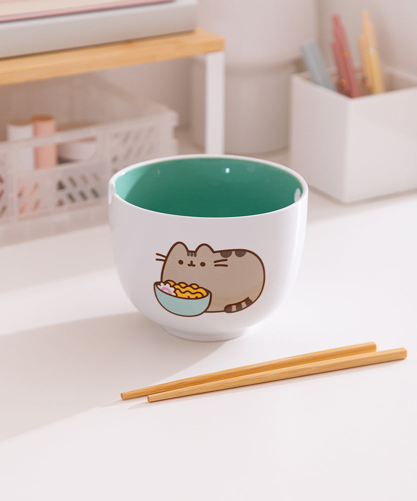 White ceramic bowl with green interior sits on a desk. The exterior features of graphic of Pusheen the Cat enjoying her own bowl of ramen.