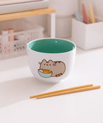 White ceramic bowl with green interior sits on a desk. The exterior features of graphic of Pusheen the Cat enjoying her own bowl of ramen.