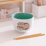 White ceramic bowl with green interior sits on a desk. The exterior features of graphic of Pusheen the Cat enjoying her own bowl of ramen.