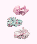 Side view of the Pusheen 3-Pack Hair Scrunchies. The trio includes light pink, mint green, and white scrunchies. Each scrunchie has a different all over print of Pusheen with various icons including hearts, stars, and more. 