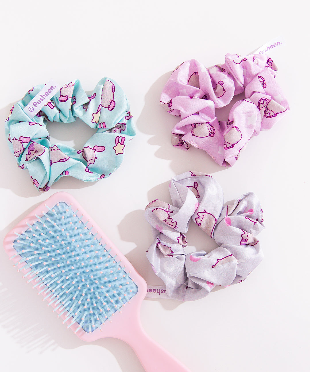 Pusheen 3-Pack Hair Scrunchies lies on a white desktop. The trio includes light pink, mint green, and white scrunchies. Each scrunchie has a different all over print of Pusheen with various icons including hearts, stars, and more. 