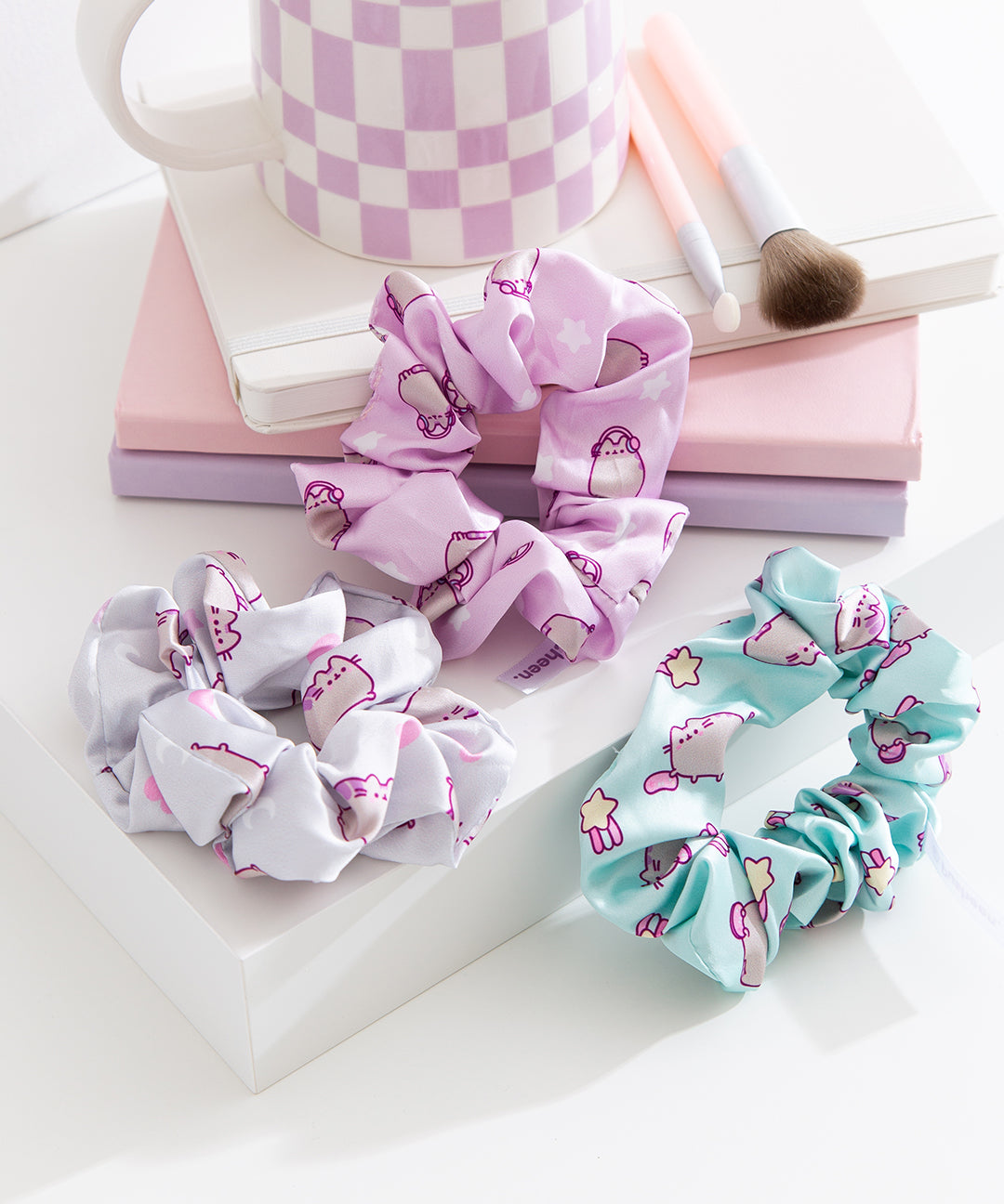 Grey, purple, and blue scrunchies lie on a desktop. The trio of hair ties feature different patterns of a pastel-colored Pusheen the Cat with heart, star, and gaming controller icons. 