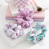 Grey, purple, and blue scrunchies lie on a desktop. The trio of hair ties feature different patterns of a pastel-colored Pusheen the Cat with heart, star, and gaming controller icons. 