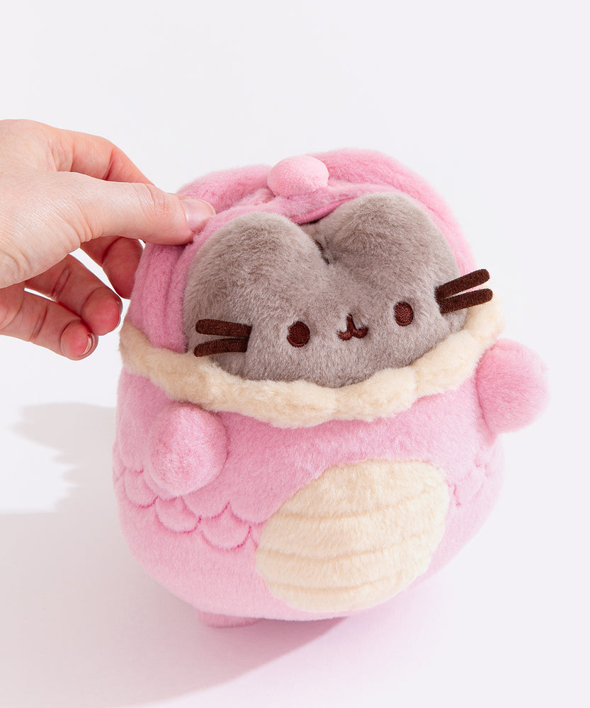 Top aerial view of a model pulling the Pink Dragon Pusheen plush’s hood off. Pusheen's fluffy grey ears and brown head stripes can be seen with the hood drawn back. 