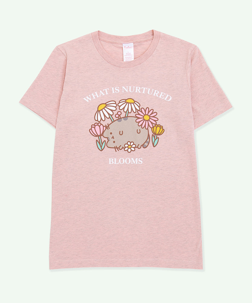 A light heather pink graphic tee against a light-green background. The center graphic features Pusheen among a variety of flowers and daisies and features the phrase “What is Nurtured Blooms” in white text above and below the Pusheen graphic. 
