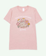 A light heather pink graphic tee against a light-green background. The center graphic features Pusheen among a variety of flowers and daisies and features the phrase “What is Nurtured Blooms” in white text above and below the Pusheen graphic. 