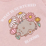 A close-up view of the Bloom Tee's screen print graphic. Pusheen lies on her back in a variety of flowers including daisies. Above and below the Pusheen graphic is the phrases “What is Nurtured Blooms.” All print elements include the colors Grey, brown, yellow, pink, green, and white. 