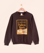 Pusheen Breads Unisex Sweatshirt hangs on a light brown hanger in front of a yellow wall. The magazine style center graphic feature Croissant, Loaf, Baguette, and Sandwich breads.