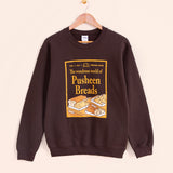 Pusheen Breads Unisex Sweatshirt hangs on a light brown hanger in front of a yellow wall. The magazine style center graphic feature Croissant, Loaf, Baguette, and Sandwich breads.