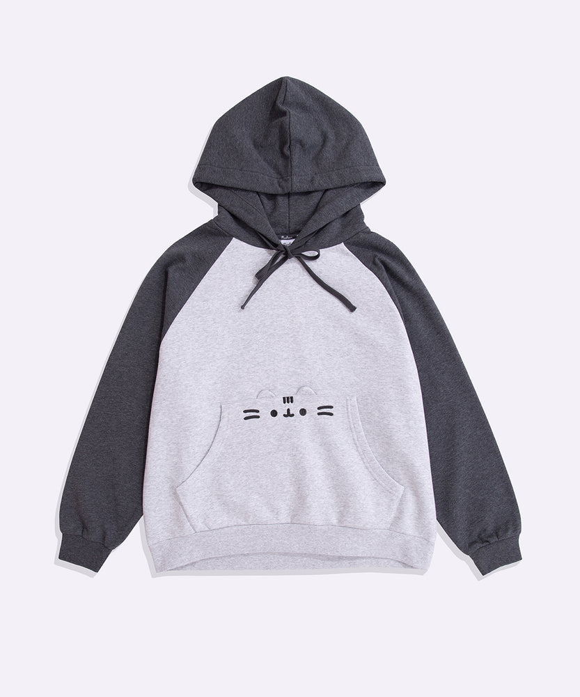 Pusheen Character Loungewear Hoodie lies on a flat surface. The two-toned sweatshirt features Pusheen’s face on the front pocket of the hoodie. The hood and sleeves are dark grey and the chest and bottom of the sweatshirt are light gray. 