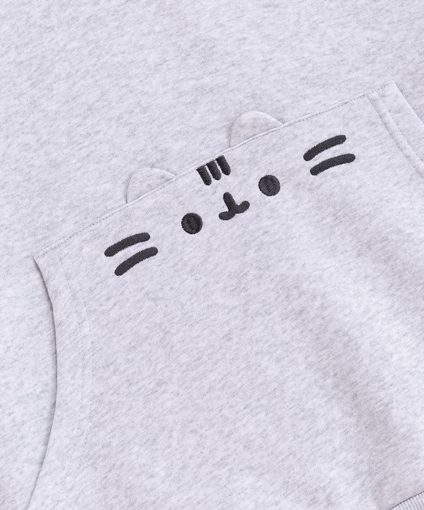 A close-up view of the embroidered graphic on the front pocket of the sweatshirt. The classic Pusheen pose shows her whiskers, mouth, eyes, head stripes in a dark gray thread. The cat’s rounded triangle ears are made of the same fabric as the hoodie and are a 3D feature of the hoodie. 