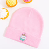 Light pink Pusheen beanie hat laid on a white surface accompanied by cupcakes with white, yellow, and pink frosting. 