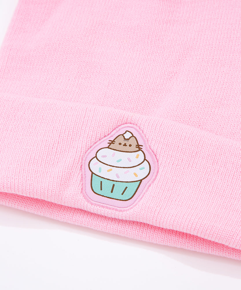 Light pink Pusheen beanie hat laid on a white surface accompanied by cupcakes with white, yellow, and pink frosting. 