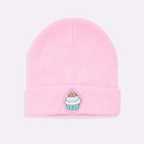 Front view of the Pusheen Cupcake Knit Hat. The light pink beanie lies on a white surface. On the front center of the folded edge is an embroidered Pusheen the Cat sitting inside the whipped icing of a cupcake. With multi-colored sprinkles and a teal base, the patch is surrounded by a light pink background. 