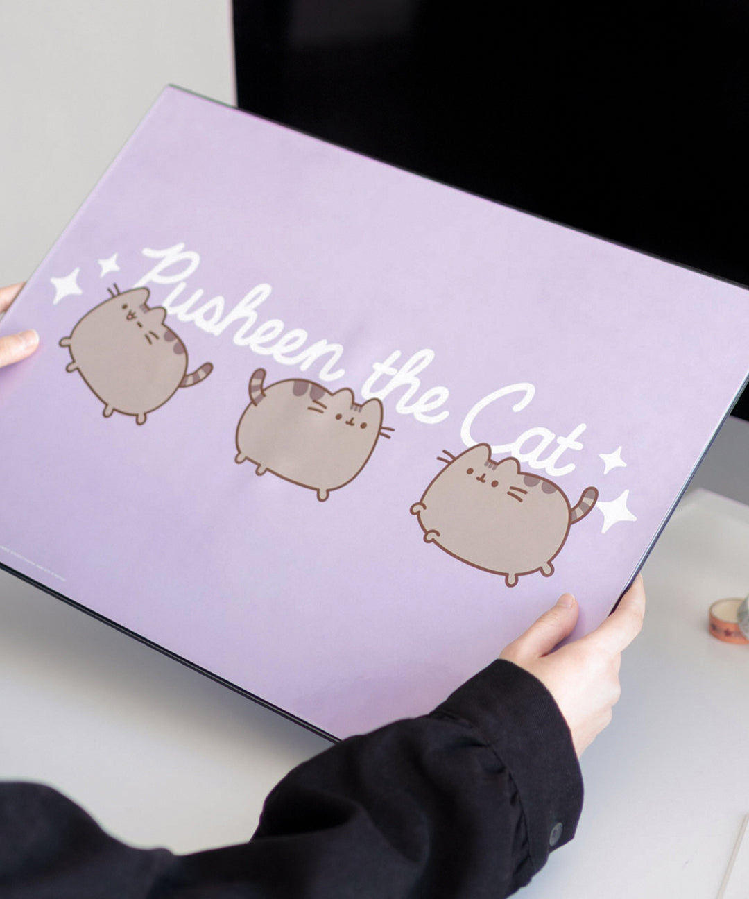 Model holds a purple rectangular desk mat. Across the middle from side to side says "Pusheen the Cat" and features three poses of the gray cat winking, walking, and leaping. 