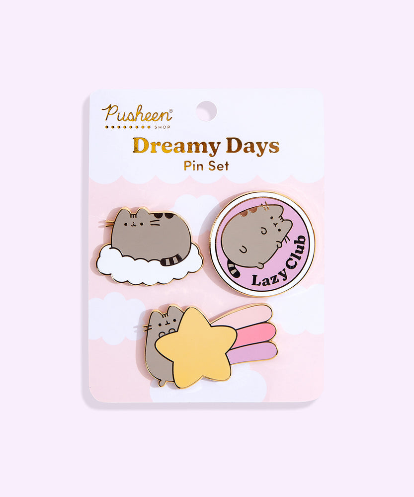 Pusheen Dreamy Days Pin Set lies in front of a light purple backdrop. Three Pusheen pins are attached to a backer card with the product name in gold foil print. The top left pin shows Pusheen resting on a fluffy white cloud. The top right pin shows Pusheen lying in a circle while the phrase “Lazy Club” is to the side of Pusheen in gold foil. The bottom pin shows Pusheen holding a shooting star with light pink, pink, and light purple streaks coming off the yellow star. 
