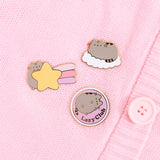 A close-up view of the three Pusheen Dreamy Days pins on a light pink cardigan. Pusheen is shown lying on a white cloud, holding a yellow shooting star, and the cat being a proud member of the Lazy Club. 