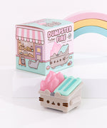 Top view of the Pusheen Dumpster Fire Vinyl Figure accompanied by its packaging box and a pastel-colored rainbow. The packaging box’s main colors are pink and mint green. On the side of the box is a bakery scene featuring Sloth, Little Sister Stormy, and delicious pastries including cakes and breads.