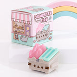 Top view of the Pusheen Dumpster Fire Vinyl Figure accompanied by its packaging box and a pastel-colored rainbow. The packaging box’s main colors are pink and mint green. On the side of the box is a bakery scene featuring Sloth, Little Sister Stormy, and delicious pastries including cakes and breads.