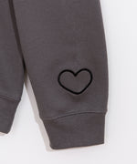 A close-up view of the embroidered graphic on the wearer’s left sleeve of the sweatshirt. The rounded heart outline is embroidered in a dark black thread on the charcoal grey sweatshirt. 