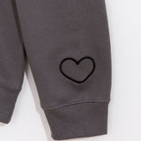 A close-up view of the embroidered graphic on the wearer’s left sleeve of the sweatshirt. The rounded heart outline is embroidered in a dark black thread on the charcoal grey sweatshirt. 