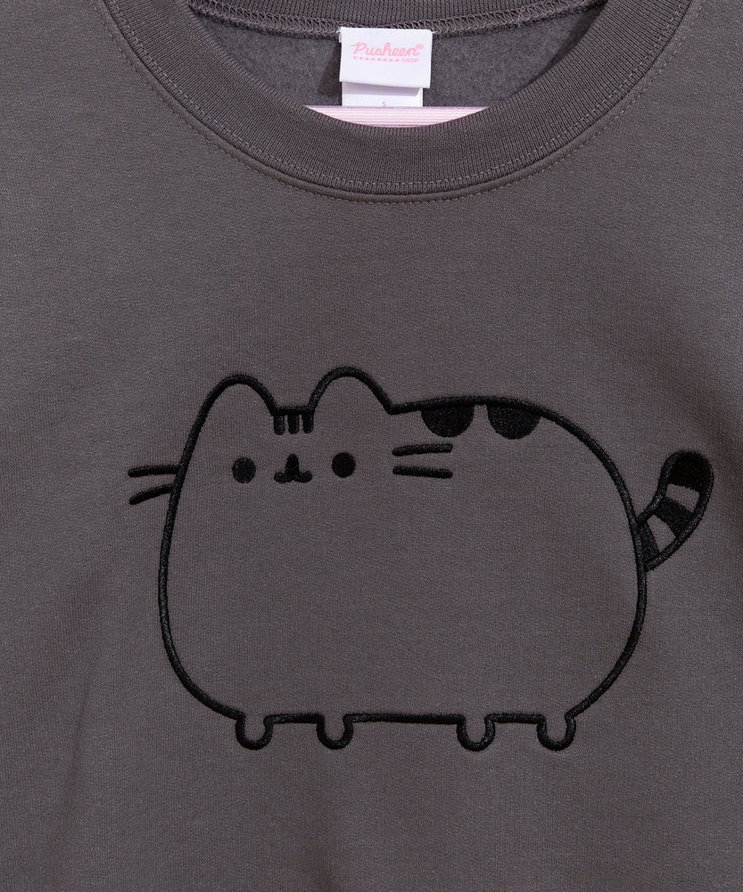 A charcoal grey sweatshirt with sleeves folded out to the side against a white background. The unisex sweatshirt has an embroidered graphic of a classic Pusheen the Cat on the center of the chest in black. The Pusheen sweatshirt also has a heart outline embroidered in black thread on the wearer’s left sleeve near the cuff. 