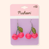 Pusheen Fruits Cherry Dangle Earrings on their cardboard backing card. The top half of the card has the Pusheen logo in brown on a light pink background and the bottom half of the card is purple. 