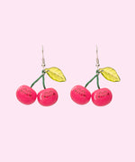 Front view of the Pusheen Fruits Cherry Dangle Earrings. Pusheen the Cat takes the form of ripe red cherries held together by a green stem and accented with a light green leaf.  