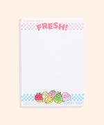 Front view of the white, rectangular Fruits notepad. At the top is a light pink checkered print with the phrases "Pusheen Fruits" and "Fresh!" At the bottom is a light blue checkered print with the phrase "Best by: Today" in light blue as well. In the center of the bottom border print are graphics of Pusheen as various fruits including a red apple, a partially peeled banana, a clapping pink strawberry, a relaxed green pear, and a winking pink and yellow peach.