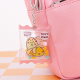 Pusheen Fruits Keyring attached to a pink backpack. The keyring dangle features a clear pouch filled with yellow, pink, and blue pom-poms. There is a graphic of Pusheen the Cat as a yellow banana on both sides with the phrase "100% Fresh"