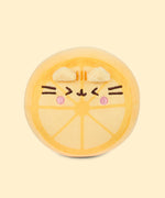 Front view of the Pusheen Fruits Lemon Squisheen Plush. The yellow plush stands 4” tall and has Pusheen the Cat’s facial features embroidered on the front of the round form in dark brown and yellow. Pusheen is blushing while making a sour face to show off the zestiness of the lemon. 