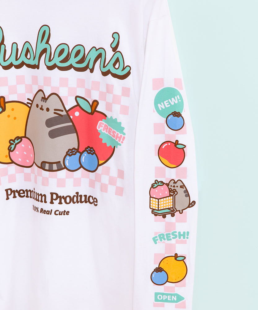 Close-up view of left sleeve of the Pusheen long-sleeve tee. The multi-color printed graphic has a light pink checkered print in the background and features the following icons: a blueberry with a “New!” sticker above, a red and yellow apple, Pusheen the Cat pushing a grocery cart with a giant pink strawberry inside, a “Fresh!” text, a blueberry and orange, and a mint green arrow sign that says “open.” 
