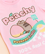 A close-up view of the Peachy Fruits Unisex Tee's screen print graphic. Pusheen sits in between two peaches with her back to the viewer and her looking over her shoulder. On the tee include the phrases “peachy,” “made with real cute,” “pusheen fruits,” “fresh,” and “100% Real Cute.” All print elements include the colors Grey, yellow, pink, green, and white. 